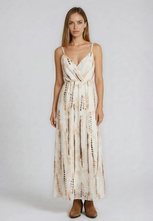 Femme portant une robe crème sans manches longue jusqu'au sol avec des rayures verticales tie-dye marron et des bottines marron, debout devant un fond uni.