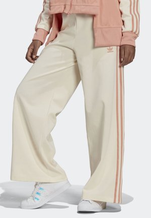 Person iført cremefarvede Adidas bukser med vide ben med pink striber og hvide sneakers, vist fra taljen til fødderne.