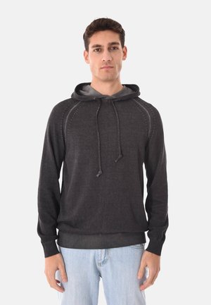 Giovane uomo con maglione grigio scuro con cappuccio e coulisse, e jeans azzurri, in piedi contro uno sfondo chiaro e uniforme.