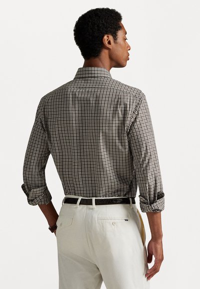 Chemise à carreaux bruns et blancs à manches longues, avec des poignets retroussés et un col classique, portée avec un pantalon de couleur claire et une ceinture sombre.