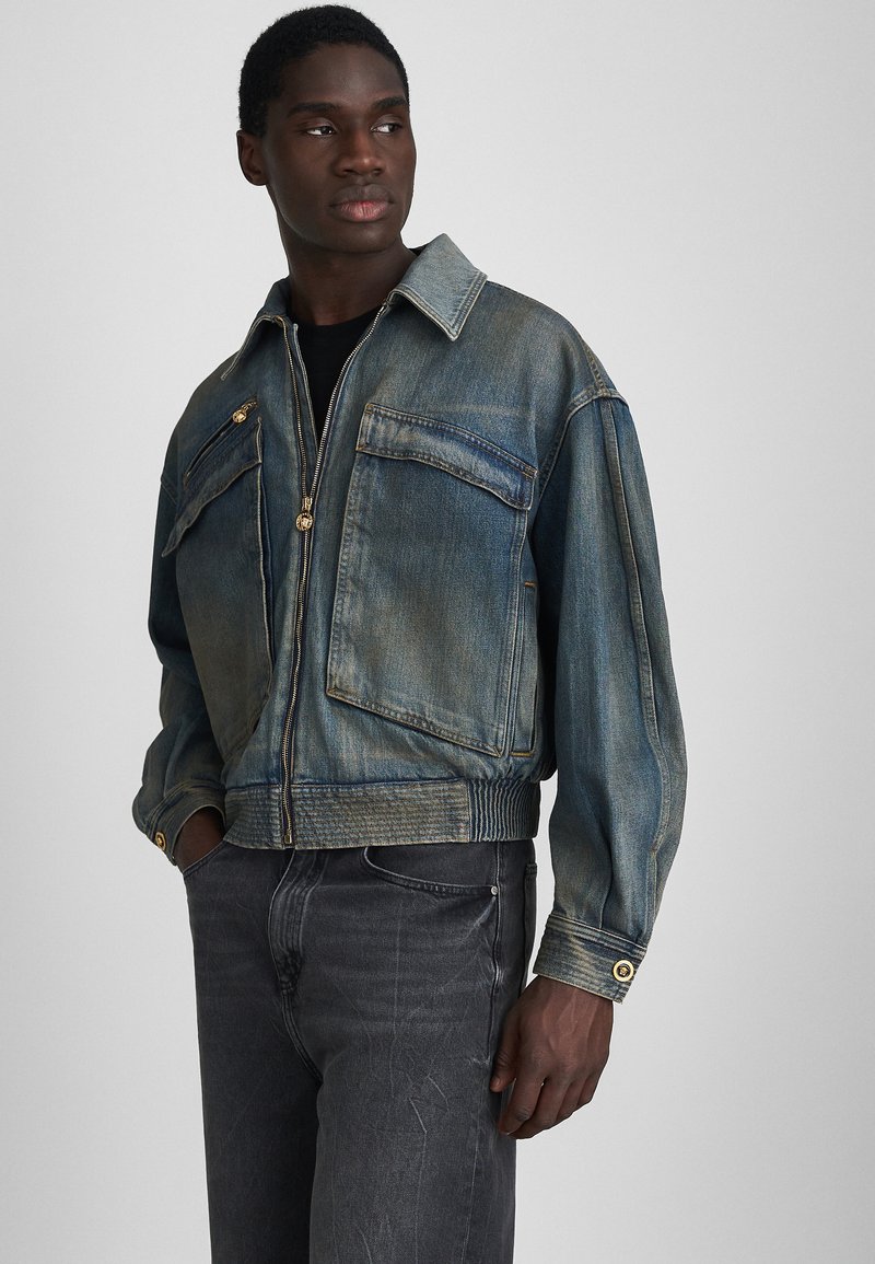 Denim jacket in faded blue featuring a front zipper, two chest pockets, ribbed hem, and gold-tone hardware accents.