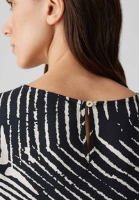 Blusa a fantasia bianco e nero, con dettaglio a chiave sul retro e chiusura con bottone, caratterizzata da una texture liscia e un design moderno.