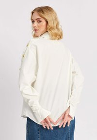 Blond naine, kandes valget nööbiga pikade varrukatega särki ja sinist teksaseelikut, seisab käed selja taga, vaadates üle õla.