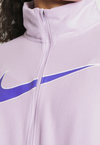 Chaqueta de cremallera en un suave tono púrpura claro con un alto cuello, textura suave y un destacado diseño del swoosh púrpura de Nike en el pecho.
