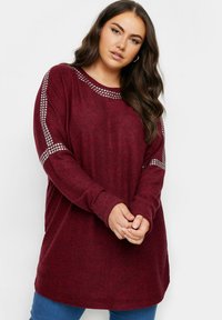 Burgundy långärmad tunika med rundad halsringning, prydda axlar med silverfärgade nitar och en mjuk, strukturerad yta.