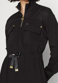 Black jumpsuit made of soft fabric, featuring a zippered collar, two front pockets, elastic waistband, and a satin ribbon tie at the waist.