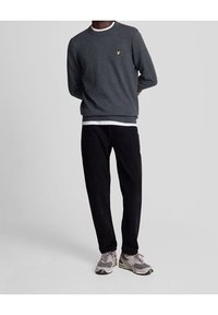 Grauer Pullover mit rundem Ausschnitt und weißem Innenkragen, schwarze Hose und graue Turnschuhe mit Akzenten. Kleines gelbes Logo auf dem Pullover.