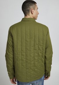 Giacca trapuntata verde oliva con colletto a punta, maniche lunghe e polsini con bottoni; presenta cuciture verticali per texture.