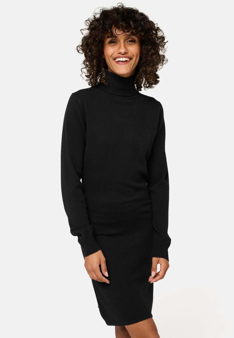 Frau mit lockigem Haar, die lächelt und ein schwarzes langärmliges Rollkragenstrickkleid vor einem schlichten hellen Hintergrund trägt.