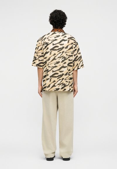 Persoon met krullend haar, gekleed in een beige en zwarte abstracte kortermige blouse en losse beige broek, staand met de rug naar de kijker.