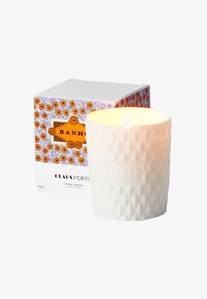 Weiße, geometrisch gemusterte Kerze in einem strukturierten Glasbehälter mit angezündetem Docht, neben einer orange-blauen Blumenschachtel mit dem Aufdruck "Claus Porto."