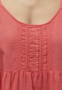 Top sin mangas color coral con escote en forma de U, que presenta pliegues y un borde de encaje decorativo a lo largo del centro. Tejido suave y texturizado con dobladillo con volantes.