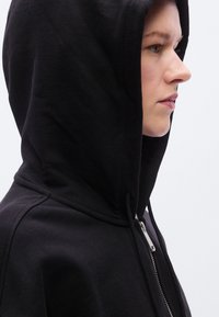 Μαύρο hoodie με φερμουάρ, μ εγάλο κουκούλι, λεία υφή και μεταλλική λεπτομέρεια φερμουάρ. Διαθέτει στενή εφαρμογή και ορατές γραμμές ραφής στο πλάι.