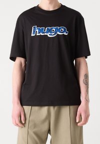 Musta puuvillainen T-paita, jossa on lyhyet hihat ja sinivalkoinen "hugo"-logo. Sileä pinta, rento istuvuus. Vaalea tausta.