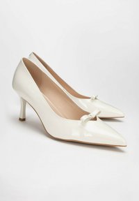 Scarpe con tacco alto in pelle patent bianca con punta affilata e un piccolo fiocco decorativo sulla parte superiore. Design elegante con tacco sottile.