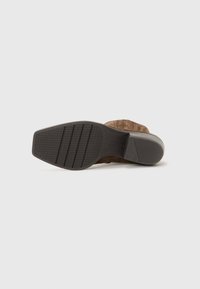 Sandalo in suede marrone con suola in gomma. La parte inferiore presenta scanalature orizzontali per una migliore aderenza e un lieve tacco. In generale, ha una texture naturale.