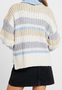 Kvinna som bär en grovstickad tröja med beige, grå, vit och blå randning, tillsammans med en svart jeanskjol.