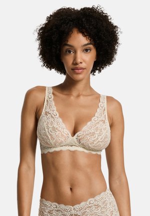 Soutien-gorge en dentelle de couleur beige clair avec un décolleté en V profond, des bords festonnés et des bretelles larges. Motifs floraux texturés sur toute la surface.