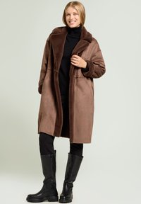 Brown faux fur coat with a wide, textured collar, paired with black leggings and knee-high black boots. Smooth fabric, oversized fit.