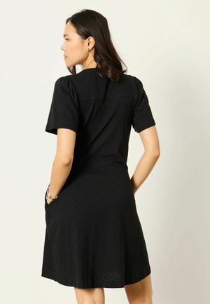 Nainen, jolla on olkapäähän ulottuvat tummat hiukset ja musta, lyhythihainen mekko, jossa on hieman puffitetut hihat, kädet taskuissa, selin katsojaan.