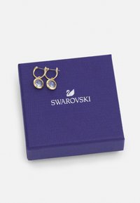 Zlatne kružne naušnice s okruglim svijetloplavim kristalima, prikazane na teksturiranoj ljubičastoj kutiji s logotipom Swarovski.