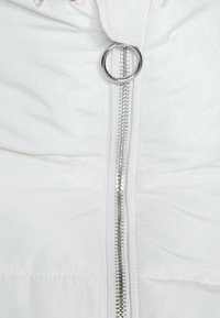 Detalle de una chaqueta de plumas blanca que presenta un cierre vertical plateado con un tirador circular y secciones cosidas que exhiben una textura acolchada.