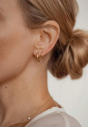 Des boucles d'oreilles en or en forme de créole sont portées dans une oreille percée, mettant en valeur une finition polie. L'arrière-plan est neutre, soulignant l'éclat des bijoux.