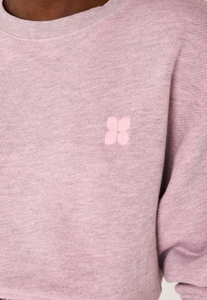 Primo piano di una persona che indossa un maglione lavorato a maglia di colore viola chiaro con un piccolo emblema a forma di fiore a quattro petali sul petto.
