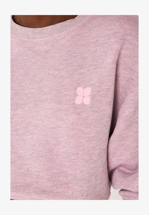 Primo piano di una persona che indossa un maglione lavorato a maglia di colore viola chiaro con un piccolo emblema a forma di fiore a quattro petali sul petto.