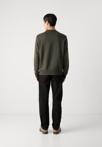 Felpa verde oliva con scollatura rotonda e polsini a costine, abbinata a pantaloni neri e scarpe beige, vista da dietro.