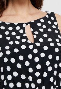 Robe noire à pois blancs, décolleté en scoop avec une ouverture en forme de goutte, tissu doux et silhouette ajustée.