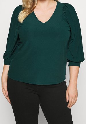 Groene V-hals top met 3/4 pofmouwen, gemaakt van zachte stof. Heeft een getailleerde lijfje en een zachte draping op de schouders. Gekleed met zwarte broek.
