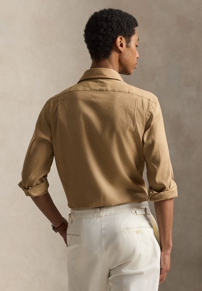 Άνδρας με σγουρά μαλλιά που φορεί καφέ πουκάμισο με γυρισμένα μανίκια και λευκό παντελόνι, στέκεται με την πλάτη του γυρισμένη προς ένα απλό μπεζ φόντο.