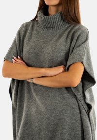 Poncho gris de punto grueso con un alto cuello de canalé, mangas cortas y detalles de botones en los laterales, que muestra una textura suave y una forma oversized.