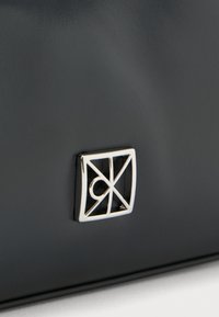 Superficie in pelle nera con un piccolo emblema quadrato di metallo che presenta linee diagonali e una forma circolare all'interno.