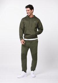 Bărbat purtând hanorac olive verde cu textură și pantaloni jogger cu genunchi nervurați asortați, cu adidași albi, stând în picioare pe un fundal alb.