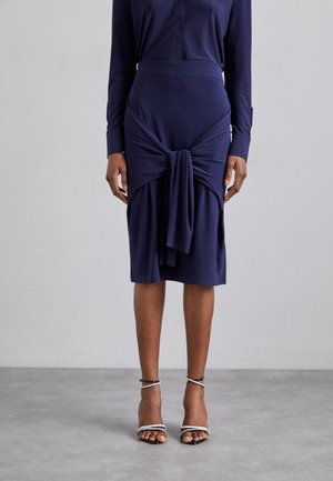 Vrouw draagt een marineblauwe rok tot op de knieën met een knoopdetail aan de voorkant en een bijpassende top met lange mouwen, gecombineerd met zwarte sandalen met bandjes.