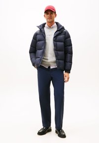 Marineblaues Pufferjackett, grauer Pullover und marineblaue Hose. Schwarze Lederschuhe. Rote Mütze. Das Model steht vor einem weißen Hintergrund.