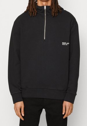Person trägt einen schwarzen Pullover mit langen Ärmeln und einem vorderen Reißverschluss sowie weißem "REPLAY Second Life"-Schriftzug auf der linken Brust.