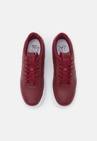 Červené kožené tenisky s hladkým povrchem, kulatou špičkou, bílou gumovou podrážkou a odpovídajícími šněrovadly. Obsahují perforace na špičce a logo na jazyku.