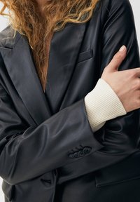 Blazer de cuero negro con solapa de muesca, bolsillo único en el pecho y puño acanalado de color crema. Textura lisa con detalles de botones en la manga.