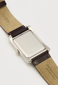 Τετράγωνη θήκη ρολογιού από ασημί ανοξείδωτο ατσάλι με καφέ δερμάτινο λουράκι. Το πάνω μέρος του δέρματος διαθέτει ανάγλυφο κείμενο· η πίσω πλευρά της θήκης είναι λεία και ανακλαστική.