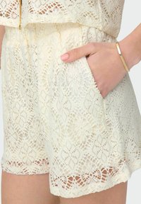 Kanten shorts in een zachte crème kleur met een bloemenpatroon, een elastische tailleband, zijzakken en een gegolfde zoom. Glad textuur zichtbaar.