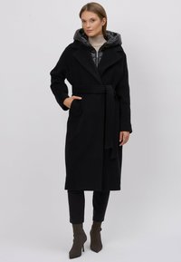 Płaszcz z czarnym paskiem i szerokim kołnierzem, noszony na pikowanej kurtce z kapturem. Strój uzupełniają dopasowane spodnie i szpiczaste botki do kostek.