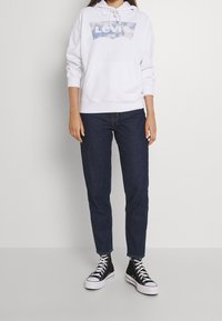 Vit hoodie med en blå Levi’s-logotyp, kombinerad med mörkblå jeans och svarta högskor. Hoodien har en dragsko i huvan.