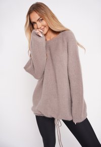Jasnobrązowy, oversize sweter wykonany z miękkiego materiału, z okrągłym dekoltem i sznurkiem ściągającym na dole. Nosi się go na czarnych legginsach.