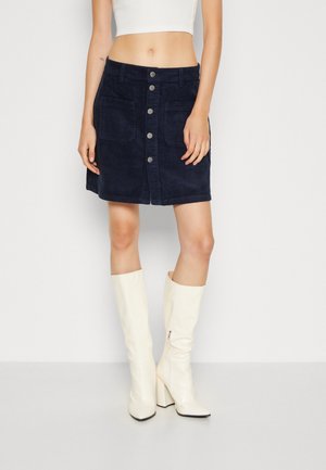 Femme portant une jupe en velours côtelé bleu marine boutonnée devant, un top blanc court, et des bottes blanches à talons hautes jusqu’aux genoux, debout devant un fond uni.