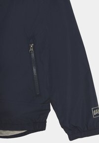 Chaqueta azul marino con un bolsillo con cremallera en el lado izquierdo, puños elásticos y una etiqueta de marca. Tejido suave y ligero con un acabado mate.