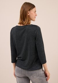 Frau mit schulterlangem Haar, die ein langärmeliges Oberteil in dunklem, gesprenkelt aussehenden Muster und graue Jeans trägt. Sie steht mit dem Rücken zur Kamera und zeigt ihr Profil.