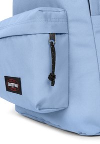 Světle modrý batoh vyrobený z odolné tkaniny, s přední kapsou na zip a černým stahovacím šňůrkou s bílými akcenty. Viditelné logo Eastpak.